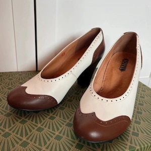 Può includere: Un paio di décolleté bicolore con tomaia color crema e punta a coda di rondine marrone scuro con perforazioni decorative brogue. Le scarpe presentano una punta arrotondata, un tacco a blocco e una fodera interna color cuoio con la scritta ROYAL.