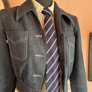 May include: A dark charcoal gray cropped jacket with white contrast stitching, two chest pockets, and a small black tag on the right pocket. Underneath is a light beige collared shirt paired with a navy blue necktie featuring diagonal red and white stripes.