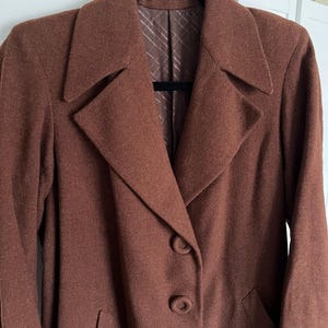 Op de afbeelding: Een bruine blazer met een klassiek ontwerp, met een reverskraag, drie knopen en twee voorzakken. De blazer hangt aan een zwarte hanger. De binnenvoering is lichtbruin met een subtiel streepjespatroon.