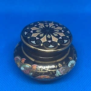 Può includere: Un piccolo vaso in ceramica nera con un coperchio dorato. Il coperchio è decorato con un motivo geometrico in oro, nero e bianco. Il vaso è decorato con una scena di persone in oro, nero, bianco e rosso.