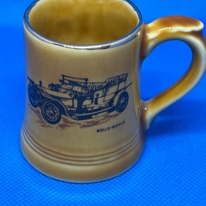 May include: A brown ceramic mug with a silver rim and a black and white image of a vintage Rolls-Royce car. The mug has the text "ROLLS-ROYCE" printed on the bottom.