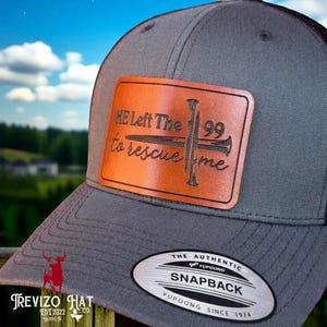 May include: Gray and black trucker hat with a brown leather patch. The patch features the text "HE Left The 99 to rescue me" with a cross and nail design. The hat has a snapback closure.