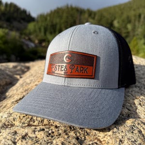May include: A gray baseball cap with a black mesh back. The front features a brown leather patch with the words "Estes Park" and the year "1917" in red lettering, along with a circular logo. The hat is sitting on a rock.