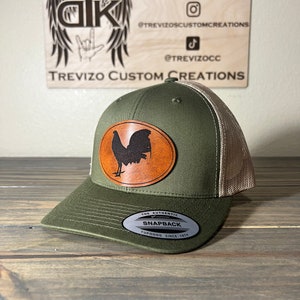 May include: A green and brown trucker hat with a brown leather patch featuring a black rooster silhouette. The hat has a mesh back and a snap closure. The hat has a label that reads "The Authentic Snapback Yupoong Since 1974".