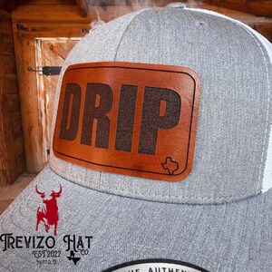 May include: A gray and white trucker hat with a brown leather patch. The patch features the word "DRIP" in bold, dark brown letters and a Texas outline. The hat has a curved brim and a mesh back.