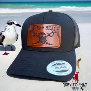 May include: A black and gray trucker hat with a brown leather patch that says "Navarre Beach" and features a skull and crossbones. The hat also has a white oval sticker that says "The Authentic Yupoong Snapback" and "Yupoong Since 1974".