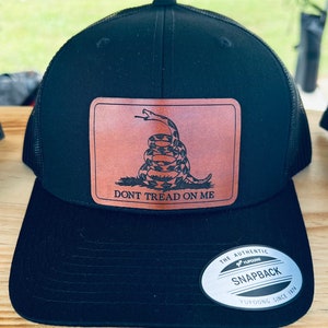 May include: A black and brown baseball cap with a leather patch featuring a coiled snake with the text "Don't Tread On Me". The cap has a mesh back and a black YUPOONG SNAPBACK label.