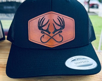 Op de afbeelding: Zwarte en bruine baseballpet met een leren patch met een paar gekruiste geweien en vishaken. De tekst "THE AUTHENTIC SNAPBACK YUPOONG SINCE 1974" staat op de achterkant van de pet.