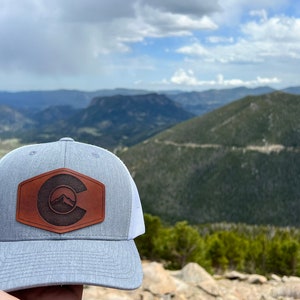 May include: A gray and white trucker hat with a brown leather patch featuring a stylized "C" with mountains inside. The hat is in front of a mountain range.