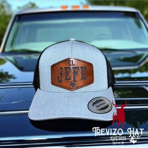May include: A gray and black trucker hat with a leather patch that reads "EL JEFE" in dark letters. The patch has a Texas outline. The hat has a snapback closure and the brand "TREVIZO HAT" is printed at the bottom.