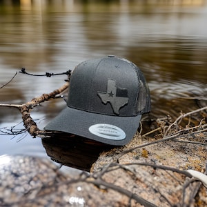 May include: A black and gray trucker hat with a black Texas state outline patch featuring a white star. The hat has a mesh back and a black adjustable strap with a silver buckle. The text "THE AUTHENTIC SNAPBACK" is printed on the buckle.