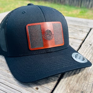 May include: Black trucker hat with a mesh back and a brown leather patch featuring the Mexican flag. The hat has a curved brim and a snapback closure. The patch displays the Mexican coat of arms.