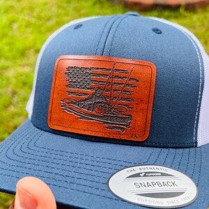 May include: A blue and white trucker hat with a brown leather patch featuring an American flag and a person fishing in a boat. The patch also includes the text "TX". The hat has a white mesh back and a "Yupoong The Authentic Snapback" label.