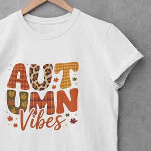 Könnte beinhalten: Weißes T-Shirt mit dem Schriftzug "AUTUMN Vibes" in einem herbstlichen Design. Die Buchstaben sind mit Karomustern, Leopardenmuster und Blättern verziert. Das T-Shirt hängt an einem Holzkleiderbügel.