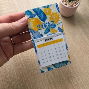 Puede incluir: Un calendario de pared 2027 con un patrón vibrante de limones amarillos y hojas tropicales azules sobre un fondo de rayas blancas y amarillas. La página del calendario muestra el mes de enero con el encabezado ENERO y una cuadrícula de fechas.