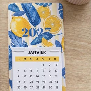 Puede incluir: Un calendario de pared 2027 con un patrón de limones y hojas azules sobre un fondo de rayas amarillas. La página del calendario muestra el mes de Janvier con los días de la semana L, M, M, J, V, S, D y fechas del 1 al 31.