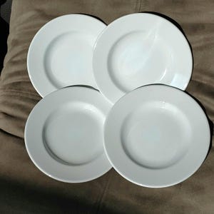 May include: Four white ceramic plates with a simple, classic design. Each plate has a slightly raised rim and a smooth, glossy surface. The plates are arranged in a square formation, perfect for a dinner setting.