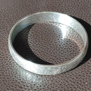 May include: A silver bangle bracelet with an engraved floral design. The bracelet is circular and has a smooth, polished surface. The engraved pattern covers the entire outer surface of the bracelet.