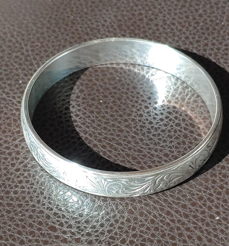 May include: A silver bangle bracelet with an engraved floral design. The bracelet is circular and has a smooth, polished surface. The engraved pattern covers the entire outer surface of the bracelet.