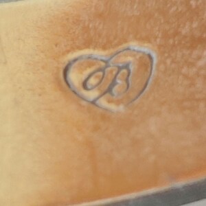 May include: Close-up of a tan leather surface with a heart-shaped embossed design. Inside the heart is a stylized letter "B". The leather has a slightly textured appearance.