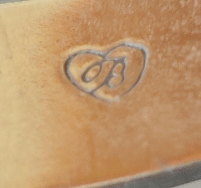 May include: Close-up of a tan leather surface with a heart-shaped embossed design. Inside the heart is a stylized letter "B". The leather has a slightly textured appearance.