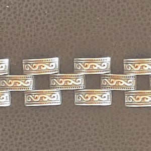 May include: Silver bracelet with rectangular links, each adorned with an engraved wave pattern. The clasp is gold-coloured. The bracelet is displayed on a brown surface. The links are approximately 1.5 cm in length.