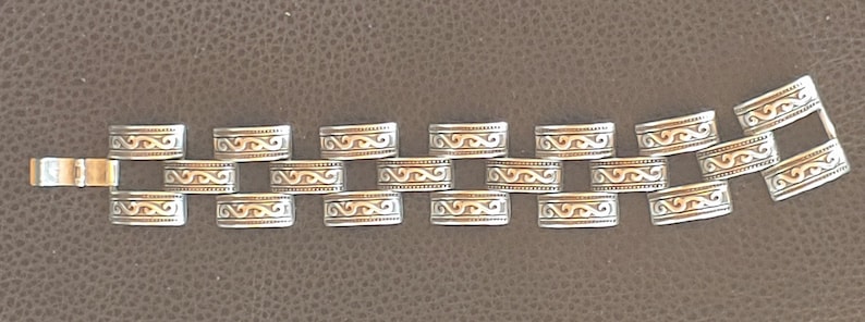 May include: Silver bracelet with rectangular links, each adorned with an engraved wave pattern. The clasp is gold-coloured. The bracelet is displayed on a brown surface. The links are approximately 1.5 cm in length.