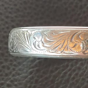 May include: Close-up of a silver engraved bracelet. The bracelet displays a detailed floral pattern with swirling leaves and delicate lines. The metal has a polished, reflective surface.