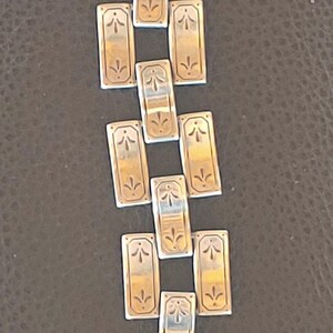 May include: A silver bracelet with rectangular links. Each link has a gold-coloured centre with a stylized floral design. The bracelet is laid out on a dark brown surface.