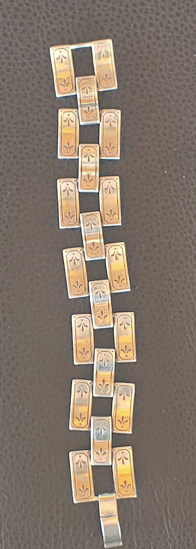 May include: A silver bracelet with rectangular links. Each link has a gold-coloured centre with a stylized floral design. The bracelet is laid out on a dark brown surface.