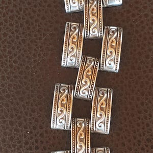 May include: Close-up of a silver bracelet with an intricate design. The rectangular links feature a repeating pattern of swirls and lines. The background is a brown, textured surface.