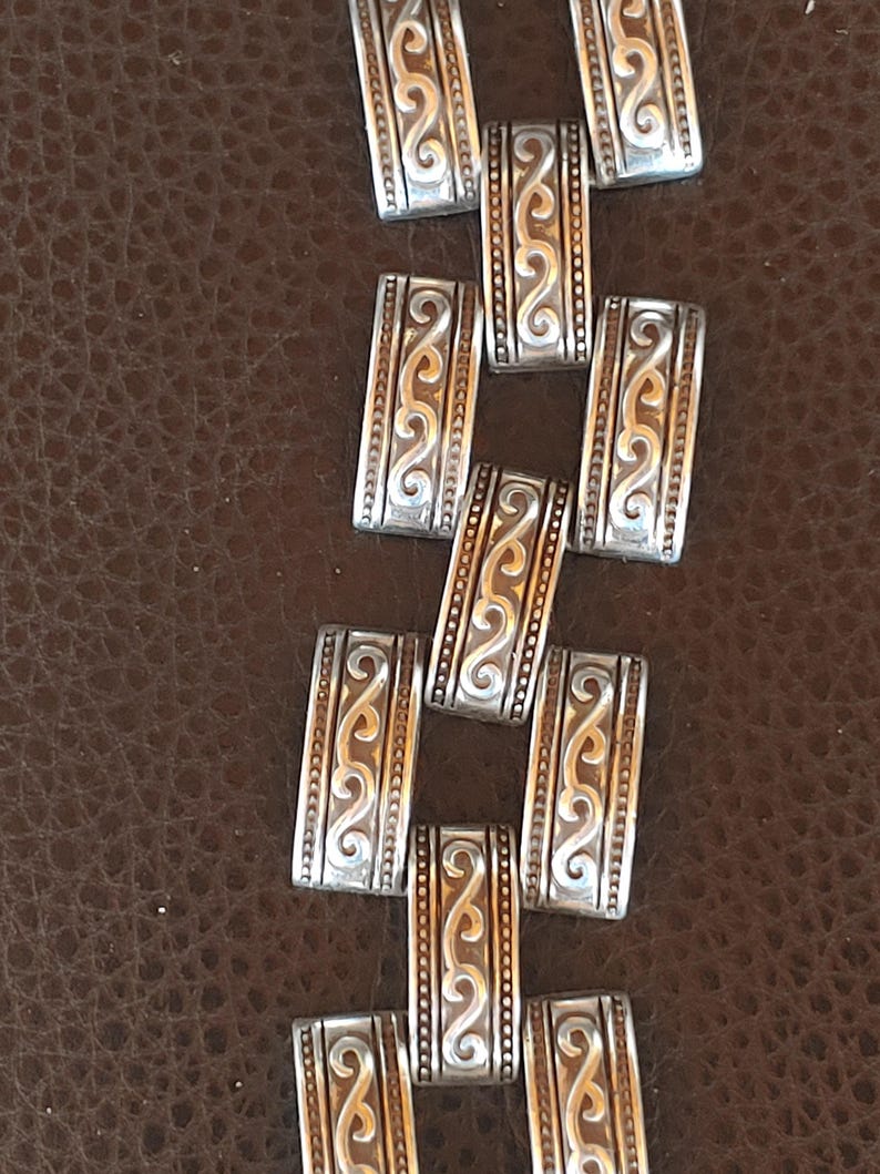 May include: Close-up of a silver bracelet with an intricate design. The rectangular links feature a repeating pattern of swirls and lines. The background is a brown, textured surface.