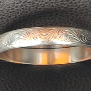 May include: A silver ring with an engraved floral pattern. The ring has a polished, reflective surface and a detailed design around the band. The ring is a piece of jewellery.