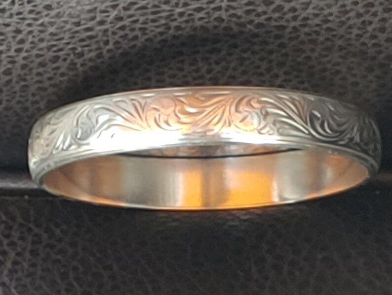 May include: A silver ring with an engraved floral pattern. The ring has a polished, reflective surface and a detailed design around the band. The ring is a piece of jewellery.