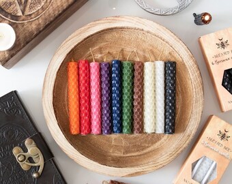 May include: A wooden bowl filled with 12 colorful beeswax candles. The candles are arranged in two rows of six. The candles are all the same size and shape, and they are all made of beeswax. The candles are a variety of colors, including red, orange, yellow, green, blue, purple, and brown. The bowl is sitting on a white surface. The text on the packaging reads "Blessed Bee - Beeswax Candles - Natural Scent".