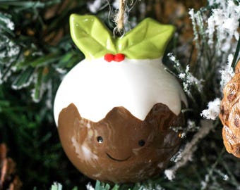 May include: A ceramic Christmas ornament shaped like a pudding, with a white top and brown base. It has a smiling face and is topped with green leaves and red berries. The ornament hangs from a twine string.