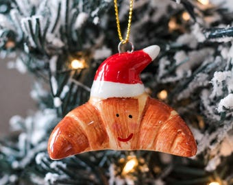 May include: A ceramic croissant ornament featuring a glossy golden-brown finish with a hand-painted smiling face and rosy cheeks. The pastry is topped with a bright red and white Santa hat and hangs from a thin gold string for display purposes.