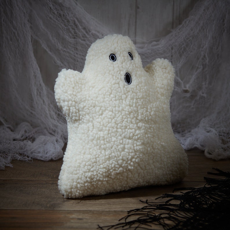 May include: A plush, white ghost-shaped pillow with embroidered black eyes and mouth. The pillow has a textured, fuzzy surface and is set against a backdrop of white fabric and a wooden surface, creating a Halloween theme.