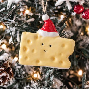 May include: A yellow Swiss cheese wedge ornament with a glossy finish, featuring a simple black dot face and a small red and white Santa hat on top. The ornament hangs from a thin, natural-coloured twine loop, designed for holiday decoration.