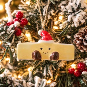 May include: A rectangular, light yellow ceramic ornament shaped like a stick of butter, featuring a small red and white Santa hat on top, two dark brown gloved hands, and a simple hand-painted face with two black eyes and a thin curved smile.