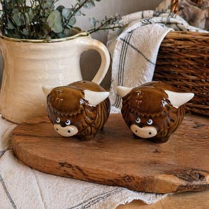 May include: A pair of brown ceramic Highland cow salt and pepper shakers with white horns and faces. They sit on a wooden board with a striped cloth. A cream jug and wicker basket are in the background.