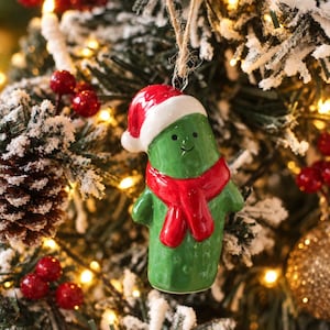 May include: A glossy green ceramic pickle ornament shaped like a character with a small smile and black dot eyes. The pickle wears a bright red Santa hat with a white brim and a matching red scarf around its neck, suspended by a thin twine loop.
