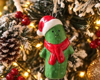 May include: A glossy green ceramic pickle ornament shaped like a character with a small smile and black dot eyes. The pickle wears a bright red Santa hat with a white brim and a matching red scarf around its neck, suspended by a thin twine loop.
