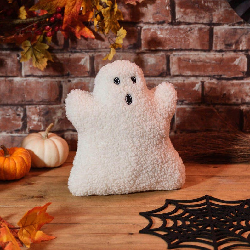 May include: A plush white ghost with black embroidered eyes and mouth, perfect for Halloween decor. The ghost is set on a wooden surface with pumpkins and a spiderweb decoration, creating a festive autumn scene.