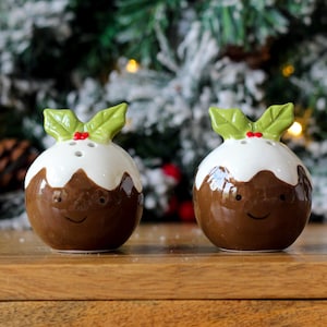 May include: A pair of ceramic Christmas pudding salt and pepper shakers. The brown pudding shakers have white frosting, green holly leaves, and red berry accents. Each has a smiling face.
