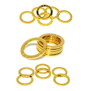 Könnte beinhalten: Goldfarbene Metallringe mit dem Text "FINA Y COCO" auf der Oberfläche eingraviert. Die Ringe sind in mehreren Stapeln und Gruppierungen vor einem weißen Hintergrund angeordnet. Die Ringe sind kreisförmig und haben ein glänzendes Finish.