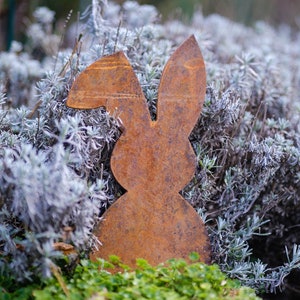 Könnte beinhalten: Ein rostiger Metall-Hasen-Ausschnitt in einer Gartenlandschaft. Der Hase ist von grünem und grauem Laub umgeben.
