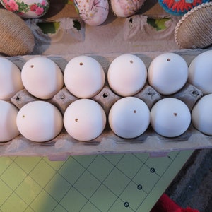 May include: A dozen white eggs with small holes in the top, presented in a cardboard egg box. Several decorated eggs are visible in the background. The eggs are a light cream colour, ideal for crafting or decoration.