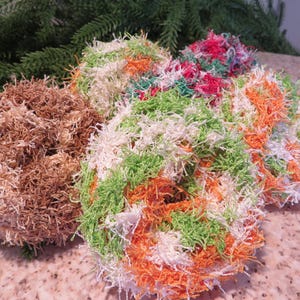 May include: A collection of handmade, textured scrubbies in various colors. The scrubbies are round and fluffy, with a mix of brown, green, orange, white, and red yarn. They are arranged on a speckled surface with evergreen branches in the background.