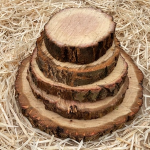 May include: A stack of four round wooden slices, each with a different diameter, arranged in a pyramid shape. The slices are made of natural wood with a brown bark and a light brown centre. The slices are sitting on a bed of straw.